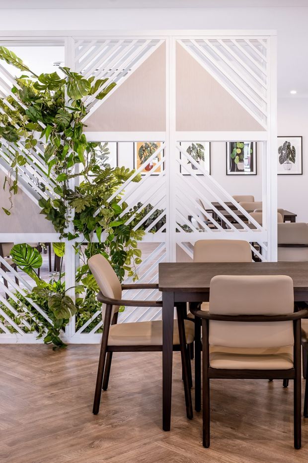 A bright dining area with a dark timber dining table and upholstered beige chairs, set against a distinctive white geometric slatted partition. Large leafy indoor plants weave through the partition, with framed botanical artwork visible beyond. The room has warm timber-look flooring, pale walls, and a clean contemporary tropical feel.