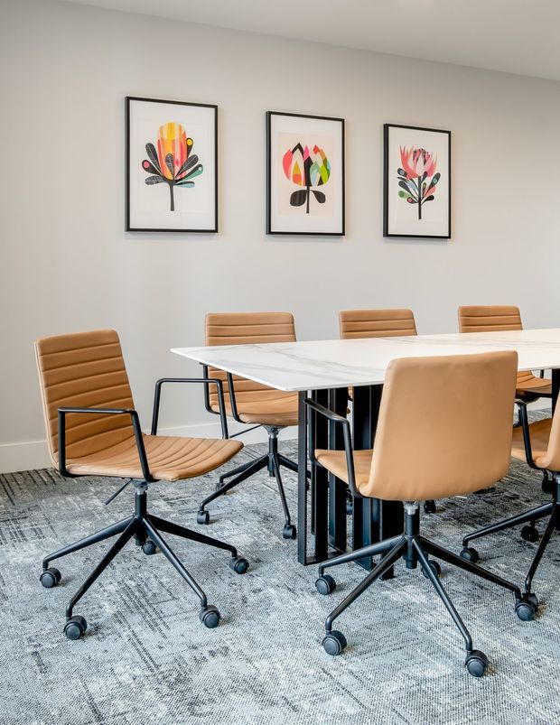 A bright contemporary dining or meeting room with a large white rectangular table surrounded by tan upholstered rolling chairs. Three colourful botanical prints in black frames hang on a light neutral wall, while a grey patterned carpet covers the floor. The space has a clean, professional and inviting atmosphere.