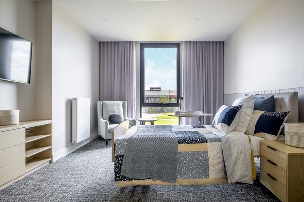 Bright, contemporary bedroom with a neatly made bed, patterned grey and navy bedding, upholstered headboard, timber bedside drawers, armchair, small round tables, full-height mauve-grey curtains, wall-mounted television, built-in low cabinetry, and a large textured carpet. A window provides an urban outlook and ample daylight.