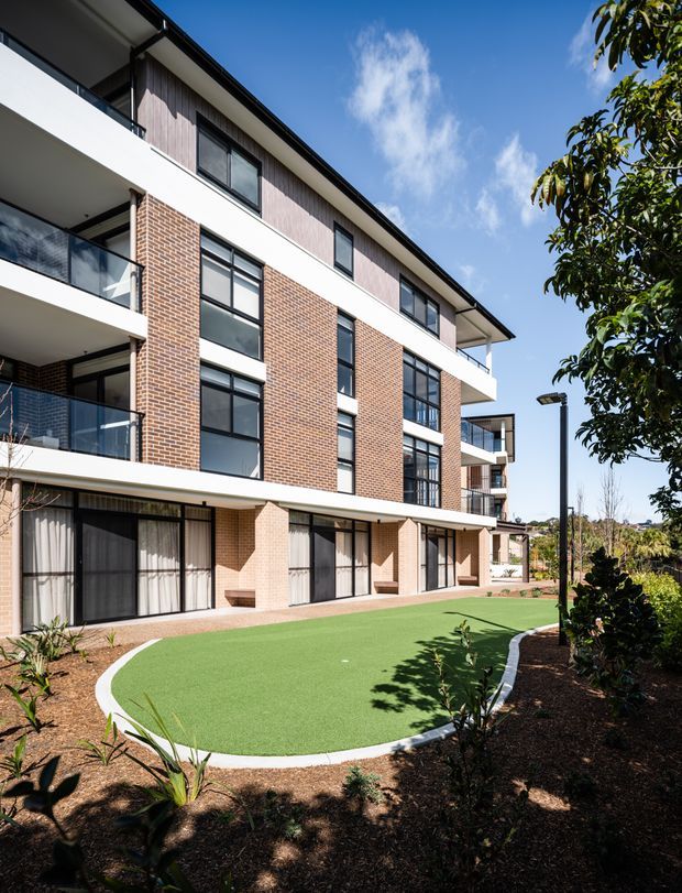 A contemporary multi-storey apartment or residential development viewed from a landscaped courtyard. The facade combines exposed red brick, pale vertical cladding, white horizontal balcony bands, dark-framed glazing, and recessed balconies with glass balustrades. A curved artificial-turf recreational area and planted garden beds occupy the foreground.