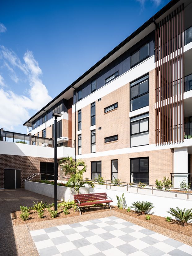 Bright exterior view of a contemporary multi-storey residential building arranged around a landscaped courtyard. The facade combines pale face brick, white rendered bands, dark window frames and upper-level dark cladding, with a prominent vertical timber-look privacy screen. The courtyard includes a paved checkerboard area, low planting, a bench, pedestrian paths and black contemporary lighting.