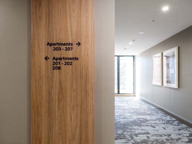 A bright modern apartment-building hallway with directional signage mounted on a vertical timber-panelled wall. The corridor has neutral painted walls, recessed ceiling lights, patterned carpet flooring, framed wall displays, and glazed doors or windows at the far end.