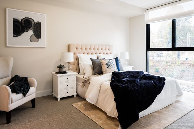 A bright, softly styled bedroom with a tufted upholstered bed, layered white bedding, navy throw, bedside chest, armchair, framed abstract artwork, and large black-framed window. The room has neutral walls, carpeted flooring, and a calm contemporary feel.