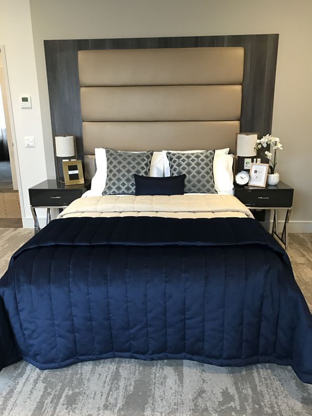 A refined bedroom centered on a large upholstered bed with a tall segmented taupe headboard framed in dark timber. Matching black bedside tables with lamps flank the bed, while patterned grey cushions, white pillows, a navy quilted coverlet, and a grey patterned rug create a polished, layered interior.