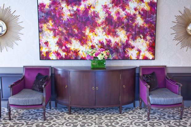 A refined interior vignette featuring a dark wood curved sideboard beneath a large vivid abstract artwork, flanked by two upholstered accent chairs in rich purple. Decorative sunburst mirrors, patterned carpet, floral wallpaper, and a small floral arrangement add layered colour and visual detail.