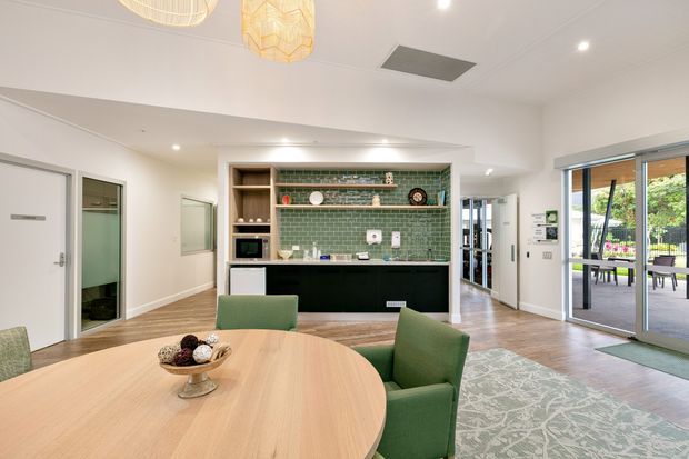 Bright open-plan dining and kitchen area with a large rounded light-wood dining table, green upholstered chairs, patterned rug, and a built-in kitchen featuring dark cabinetry, white benchtops, open timber shelving, and a green subway-tile splashback. Full-height sliding glass doors open to an outdoor patio, while a hallway and utility spaces are visible beyond.