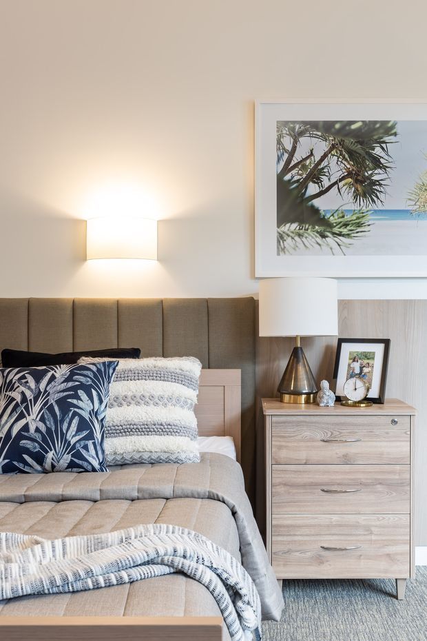 A warmly lit, softly styled bedroom featuring a tailored taupe upholstered bedhead and matching quilted bed base, layered white and grey textured bedding, and a navy botanical accent cushion. A light timber bedside chest supports a modern table lamp, clock, framed photograph, and small decor. Pale neutral walls, textured carpet, and a large framed coastal photograph create a calm coastal feel.