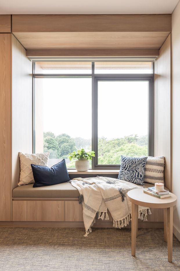 A bright built-in window seat framed by warm timber panelling and cabinetry. The upholstered bench has neutral cushions, layered patterned pillows, and a cream fringed throw, with a small round timber side table holding books and a plant. Large dark-framed glazing overlooks leafy greenery, and a textured neutral rug covers the floor.