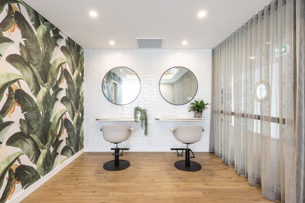 Bright contemporary salon or beauty studio with two styling stations, round mirrors, floating shelves, and adjustable salon chairs. The room features warm timber-look flooring, glossy white wall tiling, a bold tropical botanical feature wall, sheer full-height curtains, recessed lighting, and indoor plants.