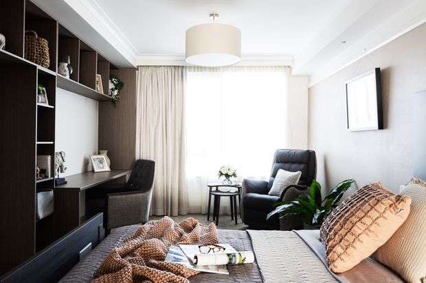 A bright, refined bedroom with a dark built-in shelving and desk unit, upholstered armchair, bedside table, large bed, layered neutral textiles, indoor plants, sheer curtains, and a contemporary pendant light. The room combines warm timber joinery with soft beige and grey furnishings.