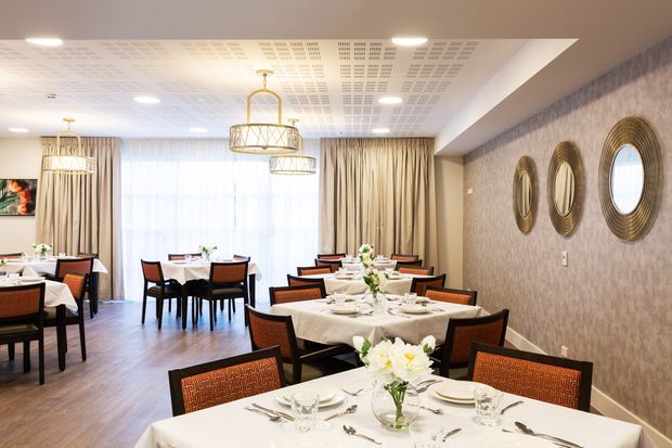 A refined dining room or restaurant interior with multiple formally set tables, dark wood flooring, upholstered dining chairs, full-height beige curtains, patterned wallpaper, decorative circular wall mirrors, and elegant pendant chandeliers. The ceiling has a perforated acoustic panel finish and recessed lighting, creating a bright, polished hospitality setting.