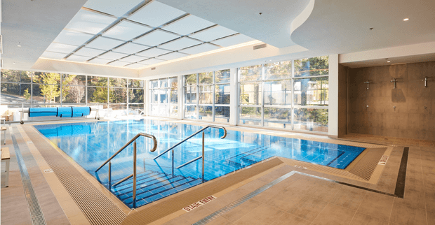 Bright indoor swimming pool area with a large tiled pool, integrated metal handrails and steps, poolside drainage grilles, and a wall-mounted shower zone. Floor-to-ceiling glazing surrounds the space, while a translucent ceiling grid and curved white ceiling detail create an airy contemporary aquatic facility.