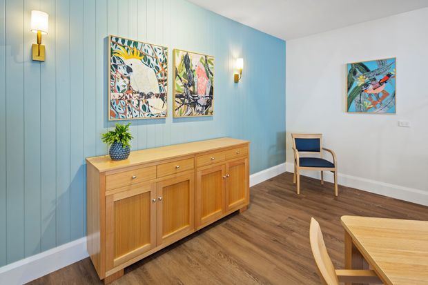 Bright, carefully styled interior showing a dining area and adjoining hallway or entry zone. The room features a warm timber sideboard with paneled doors, a matching timber dining table and chairs, blue vertical wall panelling, white walls and ceiling, timber-look flooring, decorative artwork, and contemporary wall sconces.