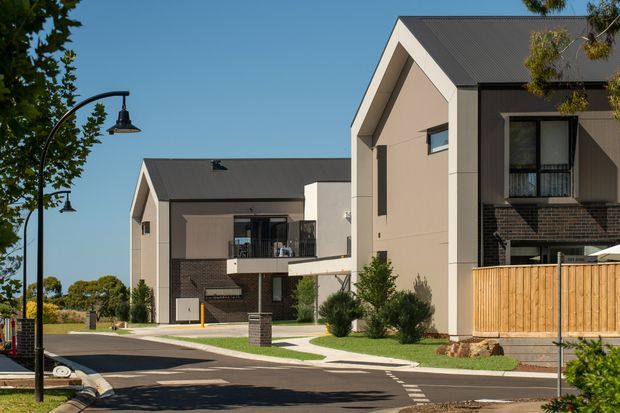 Street-level view of a contemporary multi-unit residential development with several attached or closely grouped homes. The buildings have steep gabled roofs, dark metal roofing, light rendered wall panels, dark brick lower facades, recessed balconies, black-framed windows, and timber privacy fencing. A landscaped pedestrian verge and curved black street lamps line the paved roadway.