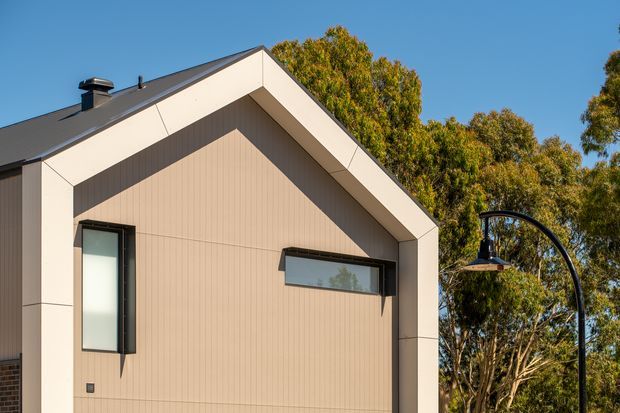 Exterior view of a contemporary single-storey gabled building facade. The facade has pale beige vertical cladding, contrasting dark window frames, a dark pitched roof, and a bold white gable surround. A curved black exterior light fitting is visible beside the building, with mature trees in the background under a clear blue sky.