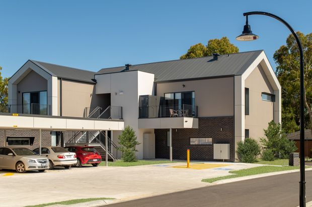 Exterior photograph of a contemporary multi-unit residential building with two prominent gabled roof forms, dark metal roofing, light fibre-cement or rendered upper façades, dark brick lower walls, balconies, external stair access and a paved parking area. A curved black street lamp is visible in the foreground, with trees and clear sky in the background.