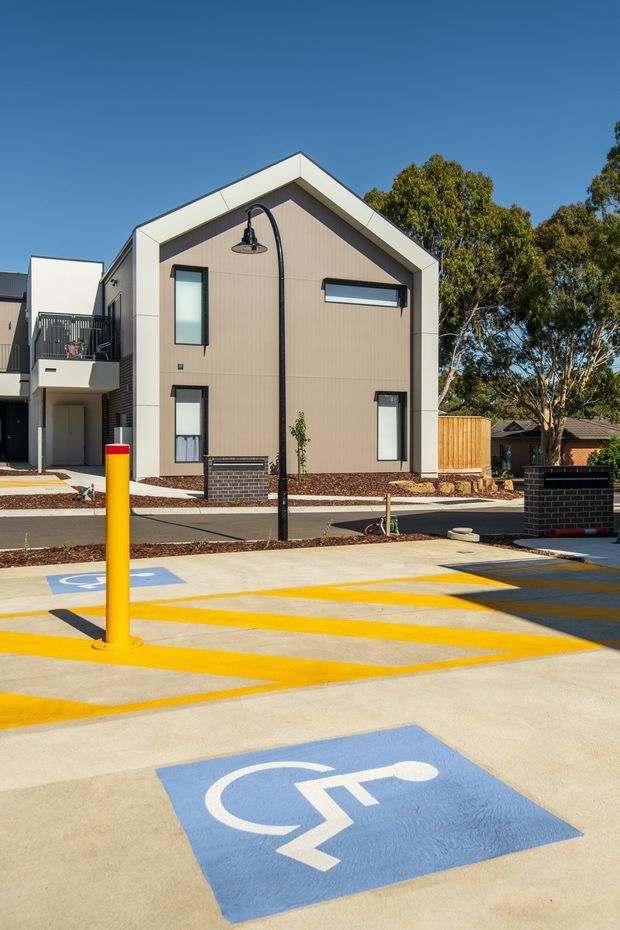 Exterior view of a contemporary multi-unit residential building with a simple gable-front form, beige vertical cladding, white fascia trim, dark-framed windows, and a small balcony. The foreground is a concrete access and parking area with bright yellow directional markings and blue accessible parking symbols. Native trees and fencing are visible around the development.