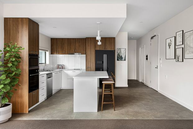 A bright open-plan kitchen and dining area with a central white island, integrated white cabinetry, dark timber wall cabinetry, stainless-steel appliances, and timber stools. The kitchen opens into a hallway or living circulation space with dark tiled flooring, white walls, recessed lighting, framed artwork, and a large indoor plant. The design is clean, contemporary, and neutral with warm timber accents.