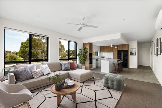 Bright open-plan living and kitchen interior with large black-framed windows overlooking greenery. A neutral sectional sofa, geometric rug, armchair and ottoman define the living area, while a white island, timber cabinetry, stainless-steel appliances and bar stools form the kitchen beyond. The space has a white ceiling with a ceiling fan, recessed lighting, framed wall art and a calm contemporary fit-out.