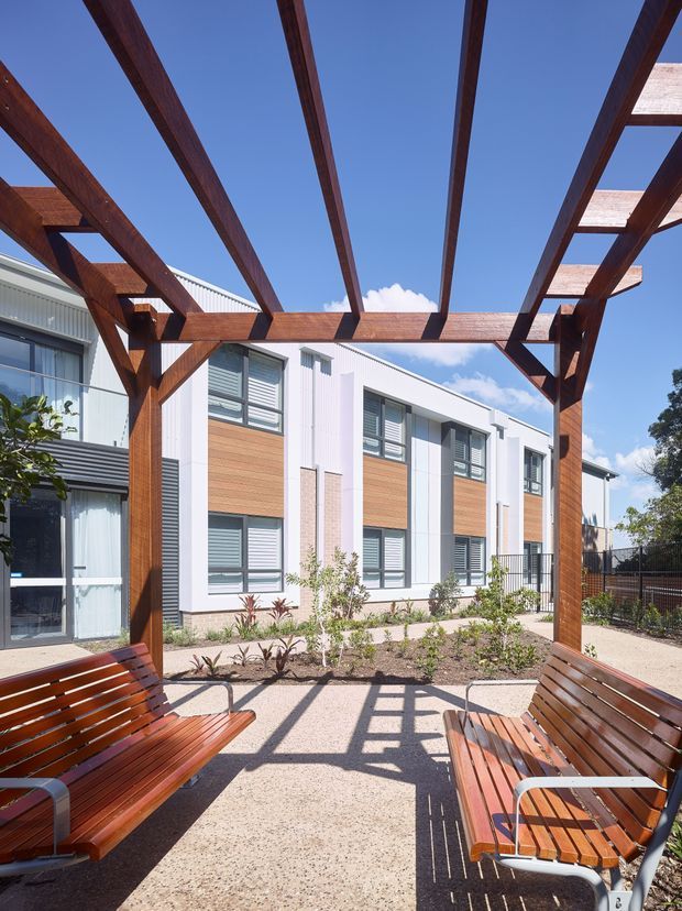 A bright landscaped courtyard framed by a substantial dark timber pergola with exposed beams and slatted timber benches. The space faces a contemporary multi-storey building with white and pale grey cladding, timber-look wall panels, extensive horizontal windows, and external louvre shading. Gravel and paved surfaces, young planting, and strong geometric shadows create a clean, sunlit communal outdoor setting.