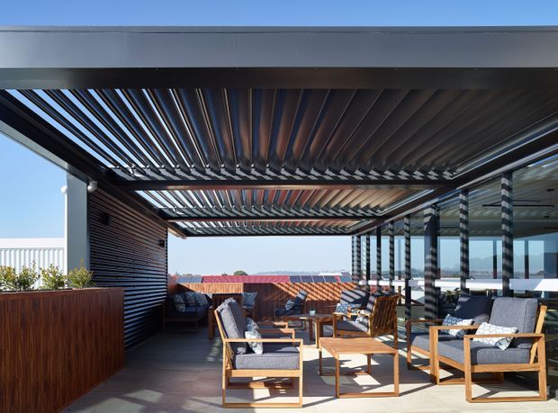 Contemporary outdoor terrace with a covered pergola featuring dark adjustable louvres, timber-lined walls, glazed screening, and timber-framed lounge seating. Built-in planter joinery and warm wood finishes create a sheltered entertaining area with strong indoor-outdoor connection.