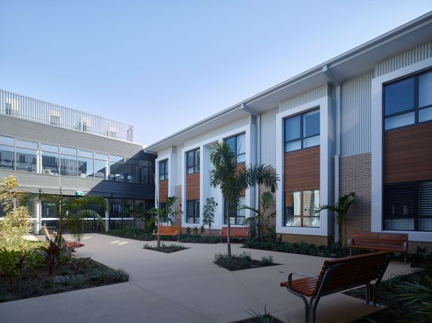 A bright contemporary multi-storey building arranged around a landscaped courtyard. The facade combines pale vertical cladding, dark metal framing, large windows, and warm timber-look wall panels. Paved paths, tropical planting, and timber benches create an inviting shared outdoor space, with a covered glazed connection visible to the left.