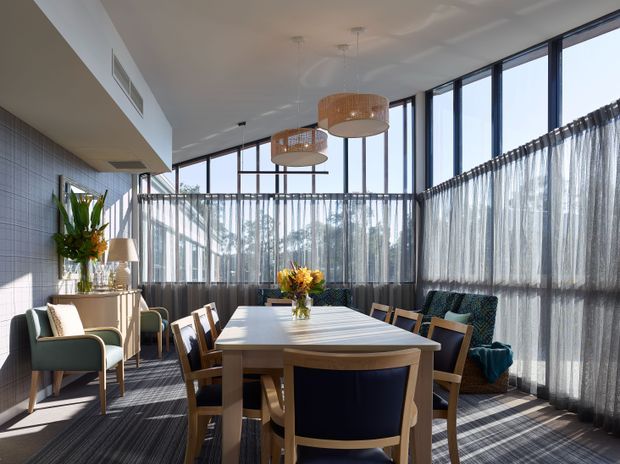 Bright, airy dining room with a large rectangular timber dining table surrounded by upholstered and timber chairs. The room features extensive floor-to-ceiling black-framed glazing with sheer curtains, a dark carpeted floor, a tiled feature wall, pendant lights with woven shades, and a small lounge seating area along the window. Natural sunlight creates strong linear shadows across the floor and furniture.