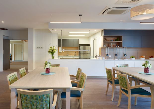 Bright contemporary communal dining and kitchen space with several light timber dining tables and colourful upholstered chairs. A large white island separates the dining area from the open kitchen, which features white cabinetry, dark blue built-in cabinetry, open shelving, pendant lighting, and a warm timber-look floor. A hallway or adjoining space is visible to the left.
