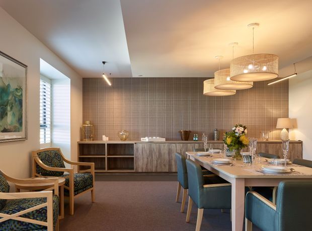 A refined open-plan dining and entertaining area with a long dining table in the foreground, upholstered blue dining chairs, and patterned timber-framed lounge chairs to the left. A timber-toned sideboard and stone-look central island sit against a full-height grid-patterned tiled feature wall. Layered pendant lights, track-style lighting, artwork, and decorative styling create a warm, contemporary interior.