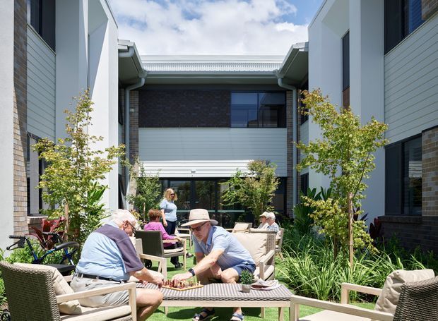A bright landscaped courtyard between contemporary multi-storey buildings, furnished with outdoor lounge seating around a low table. Residents are gathered in the central communal space, surrounded by garden planting, with a glazed entry or living area and modern cladding visible beyond.