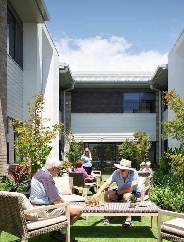 A bright landscaped courtyard between contemporary multi-storey buildings, furnished with outdoor lounge chairs and a low table where people are socialising and playing a board game. The surrounding architecture combines pale horizontal cladding, dark brick, large glazed doors and windows, and simple pitched roof forms. Planting, synthetic lawn and bicycle storage create an inviting communal outdoor space.