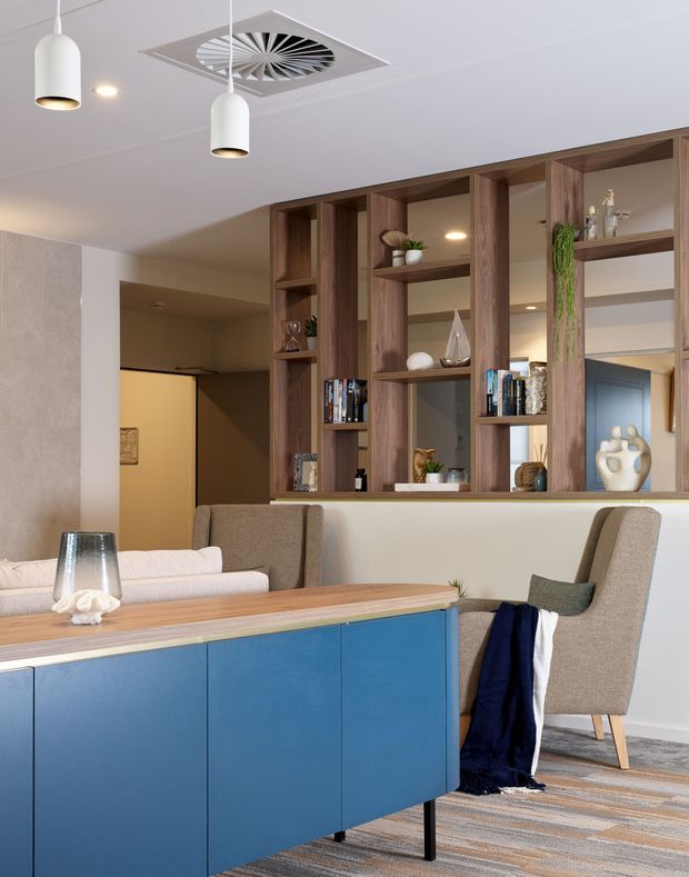 A contemporary open-plan dining and living area viewed from behind a blue built-in sideboard or island. A timber dining table is paired with upholstered dining chairs, while a full-height timber-framed open shelving divider with mirrors separates the dining space from the living area and hallway. The room has a white ceiling with pendant lights and a decorative square air-conditioning vent, pale walls, patterned carpet, and restrained decorative objects on the shelving.