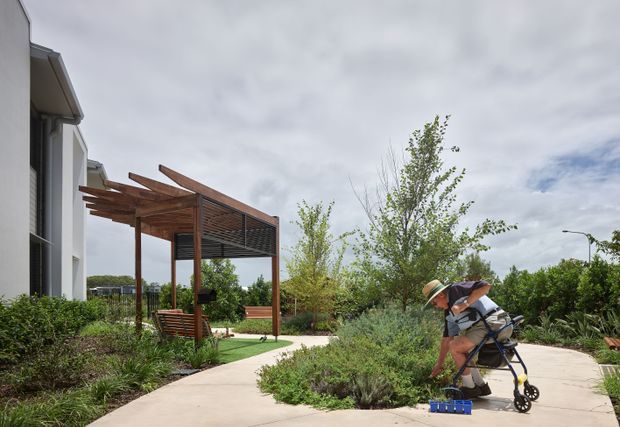 A contemporary residential exterior with a landscaped garden and pale concrete pathways. A timber pergola with exposed projecting rafters and a slatted roof creates a sheltered outdoor area beside the house. The scene includes dense planting, a bench, and a person using a mobility aid, with an overcast sky in the background.
