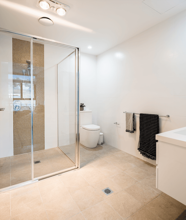 Bright contemporary bathroom featuring a large frameless-style glass shower enclosure with a small warm-toned mosaic-tile feature wall, white wall tiles, beige floor tiles, a wall-mounted white vanity, toilet, chrome fixtures, and black and grey towels.