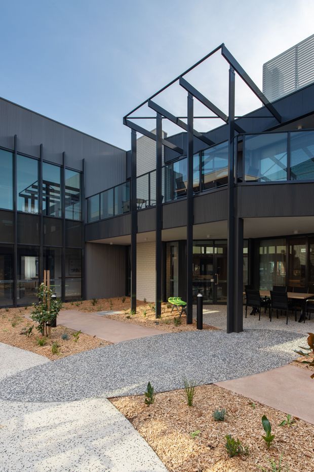 A contemporary multi-level building arranged around a landscaped courtyard. The exterior features dark metal cladding and framing, extensive glazing, exposed steel structural posts and an open pitched roof frame over a covered outdoor terrace. Gravel paths, concrete paving, sparse planting and outdoor dining furniture occupy the foreground and ground level.