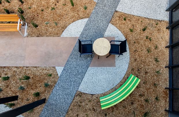 Aerial view of a designed outdoor courtyard or play area with a central circular paved zone, outdoor table and chairs, intersecting grey pathways, tan gravel landscaping, a curved green bench, and a timber seating element. Dark building edges frame the right side.