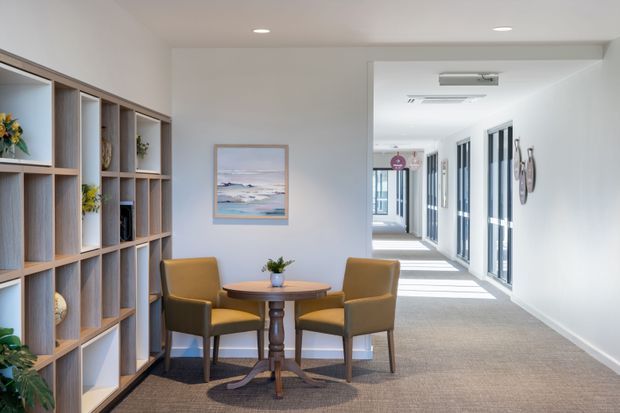 A bright, refined interior showing a small seating and reading nook beside a large built-in open shelving unit. Two mustard upholstered armchairs surround a round timber pedestal table, with a small plant and coastal artwork. A long carpeted hallway with extensive glazing and white walls extends into the background.