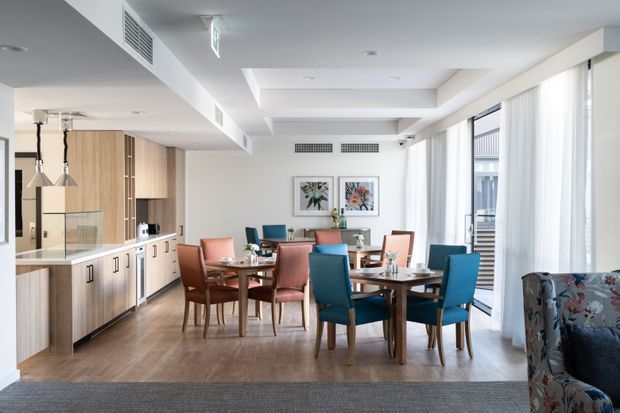 Bright open-plan interior combining a dining area and kitchen. The room features a large timber dining table surrounded by mixed upholstered chairs, extensive light timber kitchen cabinetry with a white benchtop, contemporary pendant lights, framed artwork, large windows with sheer curtains, and warm timber flooring. A patterned sofa is partially visible in the foreground.