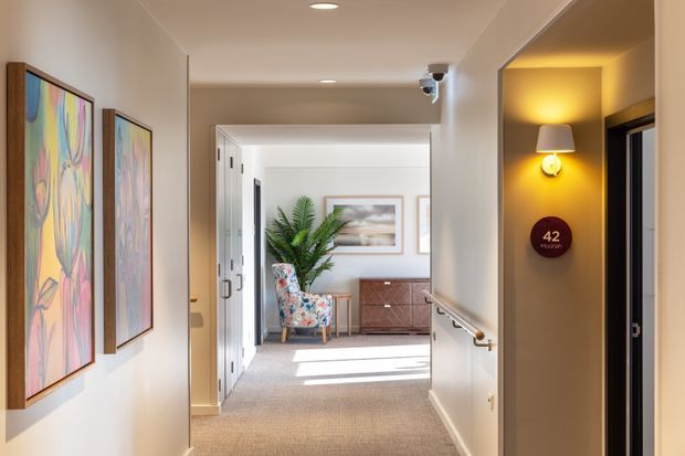 A bright, refined residential hallway viewed toward a sunlit living area. The corridor has beige painted walls, recessed ceiling lights, carpeted flooring, a timber handrail, framed colourful artwork, and a warm wall sconce beside a doorway marked “42”. At the end, a timber sideboard, large potted plant, patterned armchair, and framed artwork are visible.