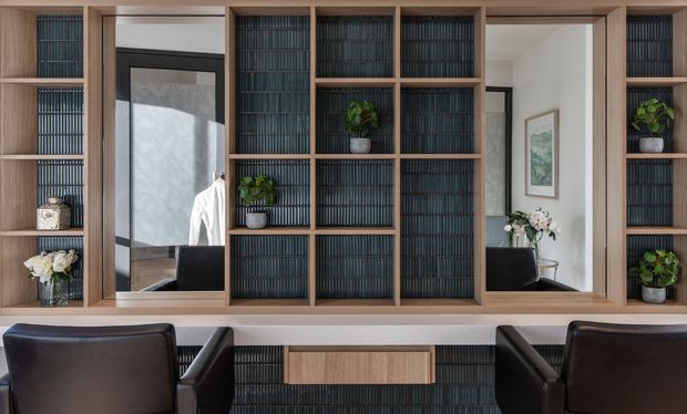 A refined built-in study or home office wall featuring warm timber shelving and cabinetry, dark vertically textured backing, and integrated open niches displaying small plants and decor. Two black leather office chairs sit at a long white work surface, with a large central mirror and a secondary reflected view of the room adding depth. The composition is symmetrical, tailored, and highly organised.