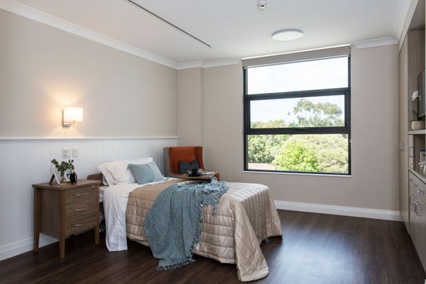 A bright, neatly styled bedroom with a single bed, dark timber flooring, pale neutral walls, white wainscoting, and a large black-framed window overlooking greenery. A timber bedside table, wall sconce, built-in shelving, and soft blue and beige bedding complete the room.
