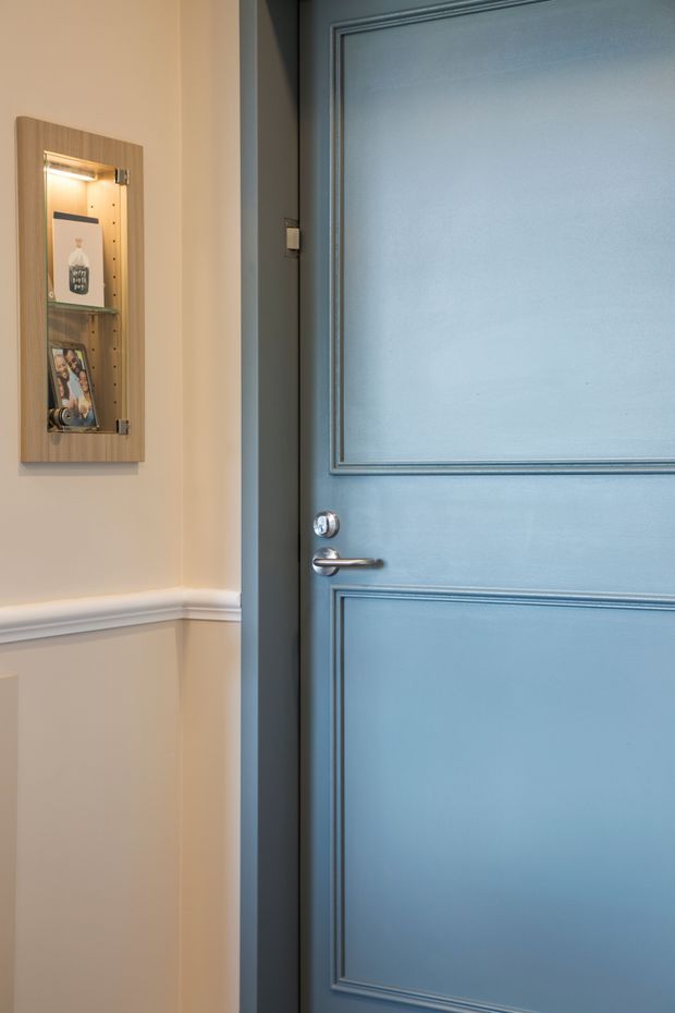 Close-up interior view of a blue painted, raised-panel door with a brushed metal lever handle and dark frame. The adjacent warm beige wall features white decorative chair-rail moulding and a slim illuminated glass display cabinet.