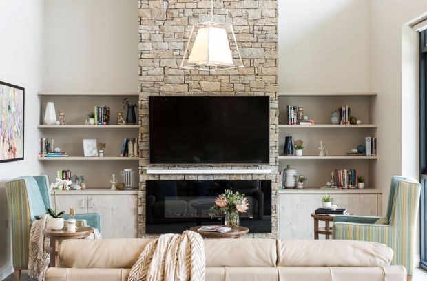 A bright, refined living room centered on a stone-clad fireplace wall with a wall-mounted television, built-in open shelving, lower cabinetry, books and decorative objects. A cream sectional sofa, striped armchairs, coffee table and large windows create a comfortable, layered sitting area.
