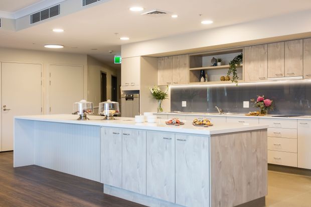 A bright contemporary commercial-style kitchen or communal kitchenette with a large light timber island, white benchtops, extensive built-in cabinetry, stainless-steel splashback, integrated appliances, open shelving, and concealed under-cabinet lighting. The space has warm wood-look flooring, recessed ceiling lights, and a clean, welcoming fit-out.
