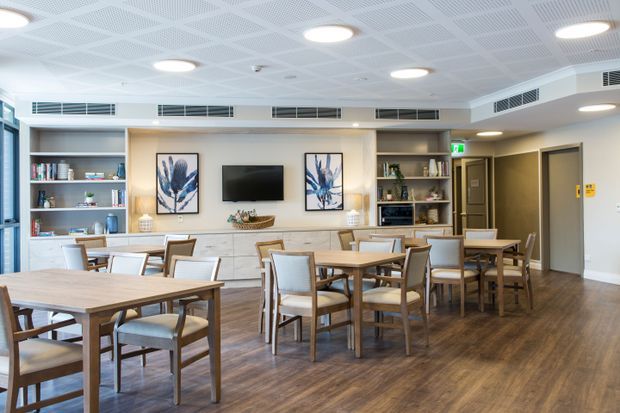 Bright communal dining and lounge area with multiple timber dining tables and upholstered chairs, built-in white cabinetry and open shelving, artwork, a television, and a small kitchenette or refreshment station. The room has warm wood-look flooring, neutral walls, and a suspended acoustic ceiling with recessed lighting and linear air-conditioning vents.