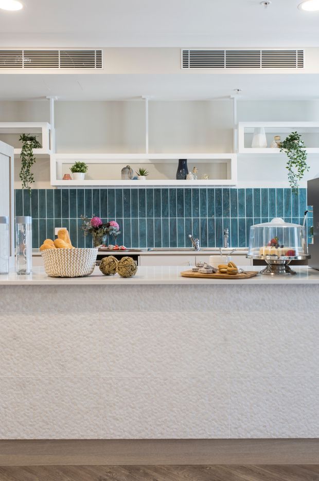 Bright contemporary kitchen with a large white textured island, deep blue vertical ceramic tile splashback, white open shelving and cabinetry, integrated ventilation grilles, and light timber flooring. Decorative styling includes plants, pottery, food displays, and a glass cake dome.