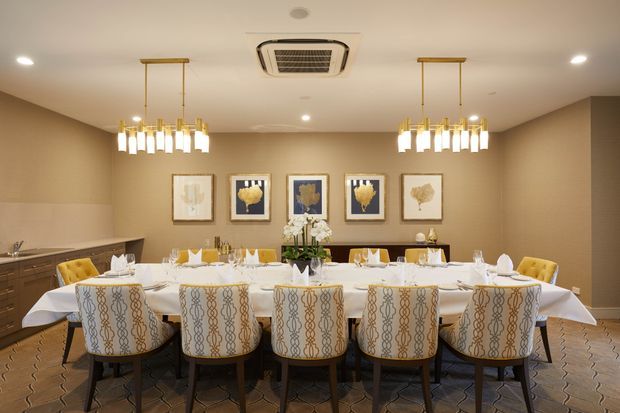 A formal dining room with a long table set for a large gathering, upholstered patterned dining chairs, warm beige walls, decorative artwork, a sideboard, and two contemporary chandelier fixtures. A compact bar or kitchenette area is visible at the left.