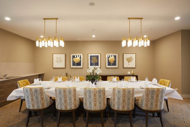A formal dining room with a long, fully set dining table surrounded by upholstered patterned chairs. The space features warm beige walls, a patterned carpet, dark wood cabinetry and sideboard, framed botanical artwork, and two contemporary brass-toned pendant chandeliers.