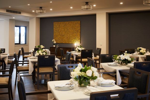 A refined contemporary dining room or restaurant interior with multiple formally set tables, dark upholstered dining chairs, white tablecloths, floral centrepieces, and warm ambient lighting. Dark charcoal feature walls frame a large textured gold artwork, while timber flooring, recessed ceiling lights, and modern pendant fixtures create an elegant atmosphere.