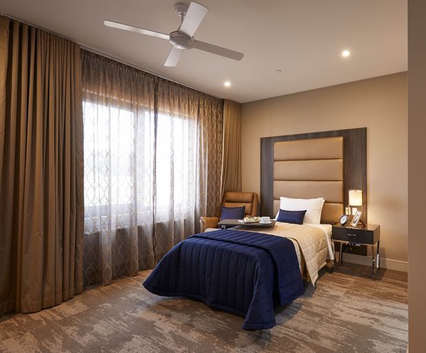 A refined bedroom with a centrally placed upholstered bed, tall padded headboard framed in dark wood, navy quilt and cushions, and matching bedside tables. Full-height layered curtains cover a large window, while recessed ceiling lights and a contemporary ceiling fan provide illumination and ventilation. The room has a warm neutral palette with patterned carpet and textured drapery.